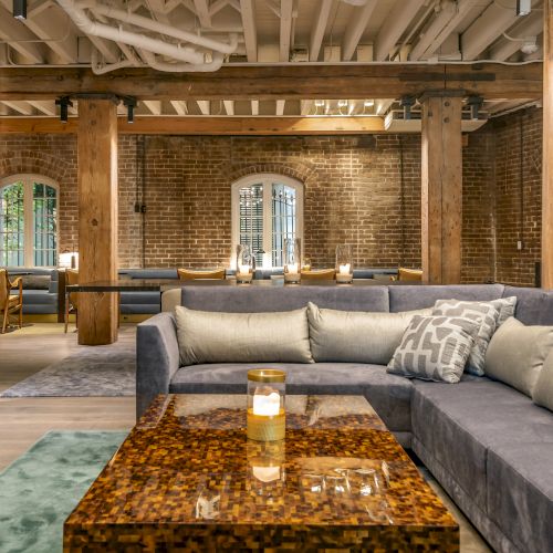 A modern and cozy lounge with wooden beams, exposed brick, a grey sectional sofa, and a coffee table with candles, providing an inviting atmosphere.