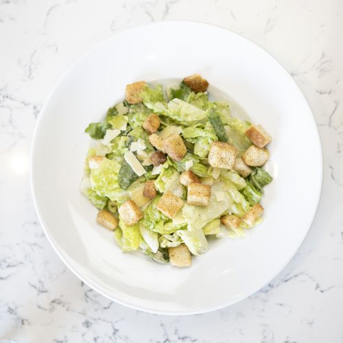 A white plate with a Caesar salad topped with croutons, placed on a white marble surface.