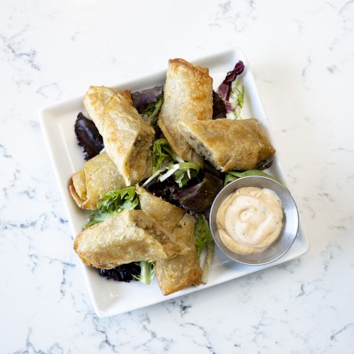 The image shows a plate of stuffed pastry rolls on a bed of greens, accompanied by a small container of dip on the side.