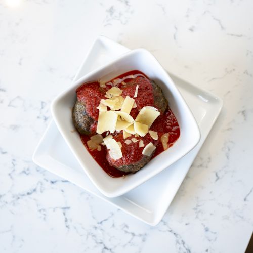 A dish of meatballs topped with tomato sauce and shaved cheese, served in a white square bowl.