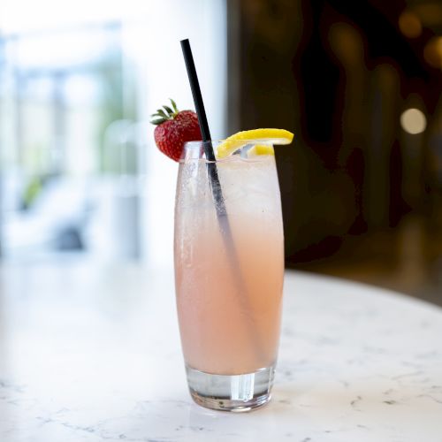 The image shows a tall glass with a pink drink, garnished with a strawberry and a lemon slice, and a black straw, on a white marble table.