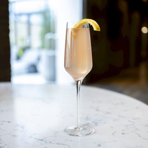 A tall, slender glass filled with a light-colored beverage, garnished with a lemon twist, sits on a marble table in a softly lit setting.