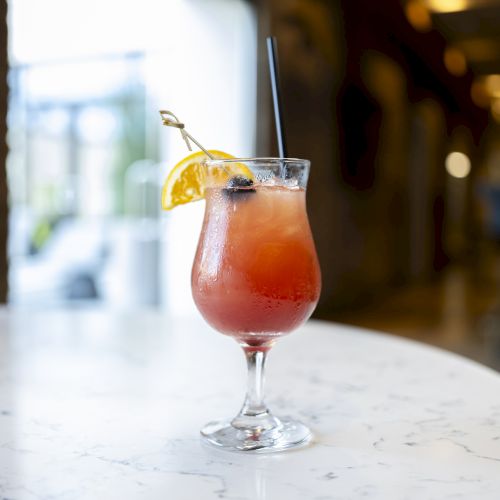 A refreshing cocktail in a tall glass with a slice of lemon, a cherry, and a straw, placed on a marble table indoors.