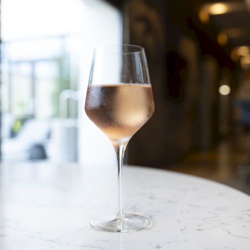 A glass of chilled rosé wine sits on a marble table surface in an elegant indoor setting.