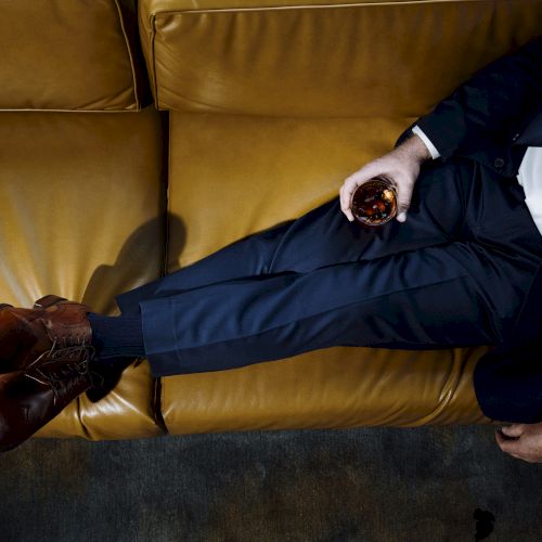 A person in a suit is sitting on a brown leather couch, holding a glass of dark liquid in their right hand and crossing their legs.