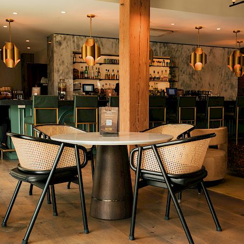 The image shows a modern cafe interior with a table and chairs in the foreground, and a stylish bar in the background adorned with hanging lights.