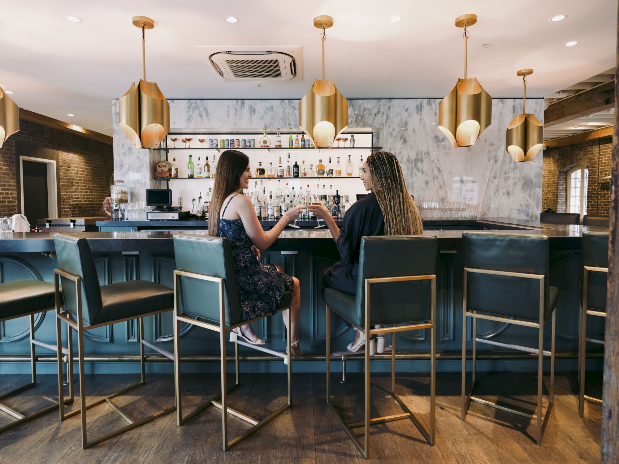 Two people sit at a stylish bar with gold pendant lights, chatting while facing a well-stocked shelf of drinks.