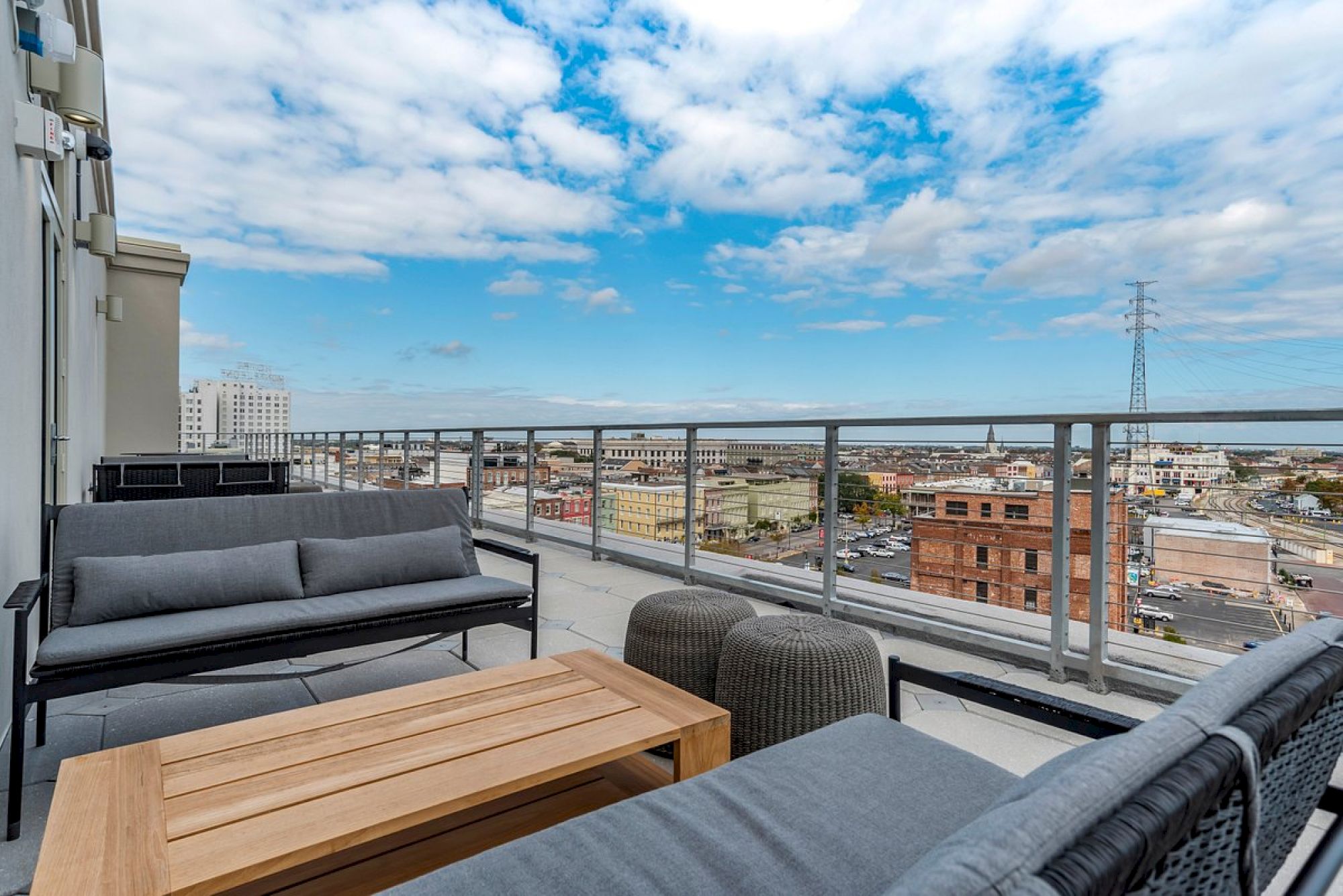 A rooftop terrace with outdoor seating, including sofas and a wooden coffee table, overlooks a cityscape under a partially cloudy sky.