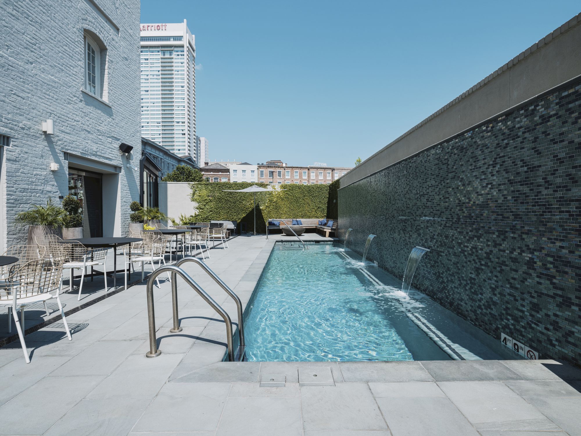 A patio with a narrow swimming pool beside a wall, surrounded by chairs and tables, and a tall building in the background.