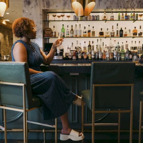 A person is sitting at a bar counter with various bottles in the background, wearing a blue dress and white shoes, pointing at something.