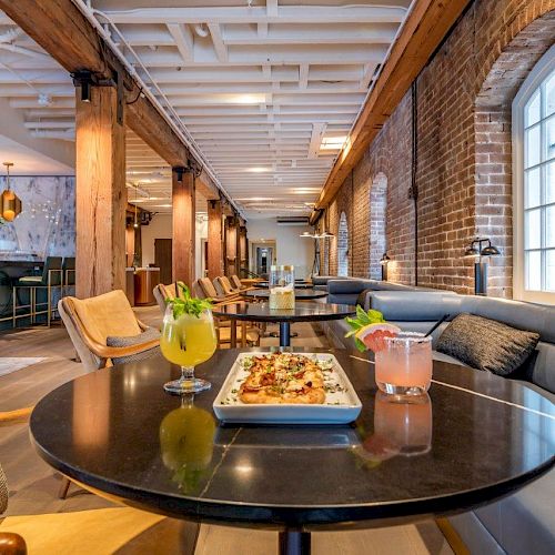 A cozy restaurant with brick walls, modern seating, a bar in the background, and a table set with colorful drinks and a plate of food in the foreground.