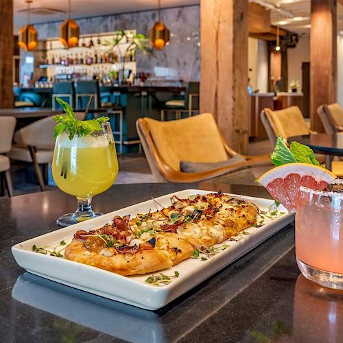 A restaurant setting with a stylish interior, featuring flatbread on a plate alongside two cocktails garnished with fruit and herbs, on a table.