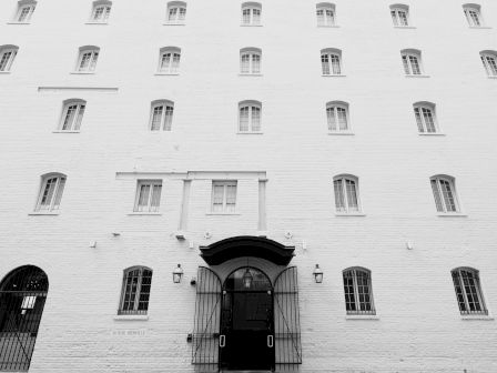 A black-and-white image shows a multi-story building with numerous small windows and a main entrance featuring iron gates and an arched canopy.