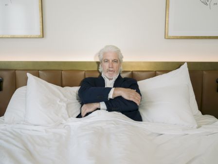 An older man with white hair and beard sits upright in a bed with a brown headboard, surrounded by white pillows and bedding, wearing a dark suit.