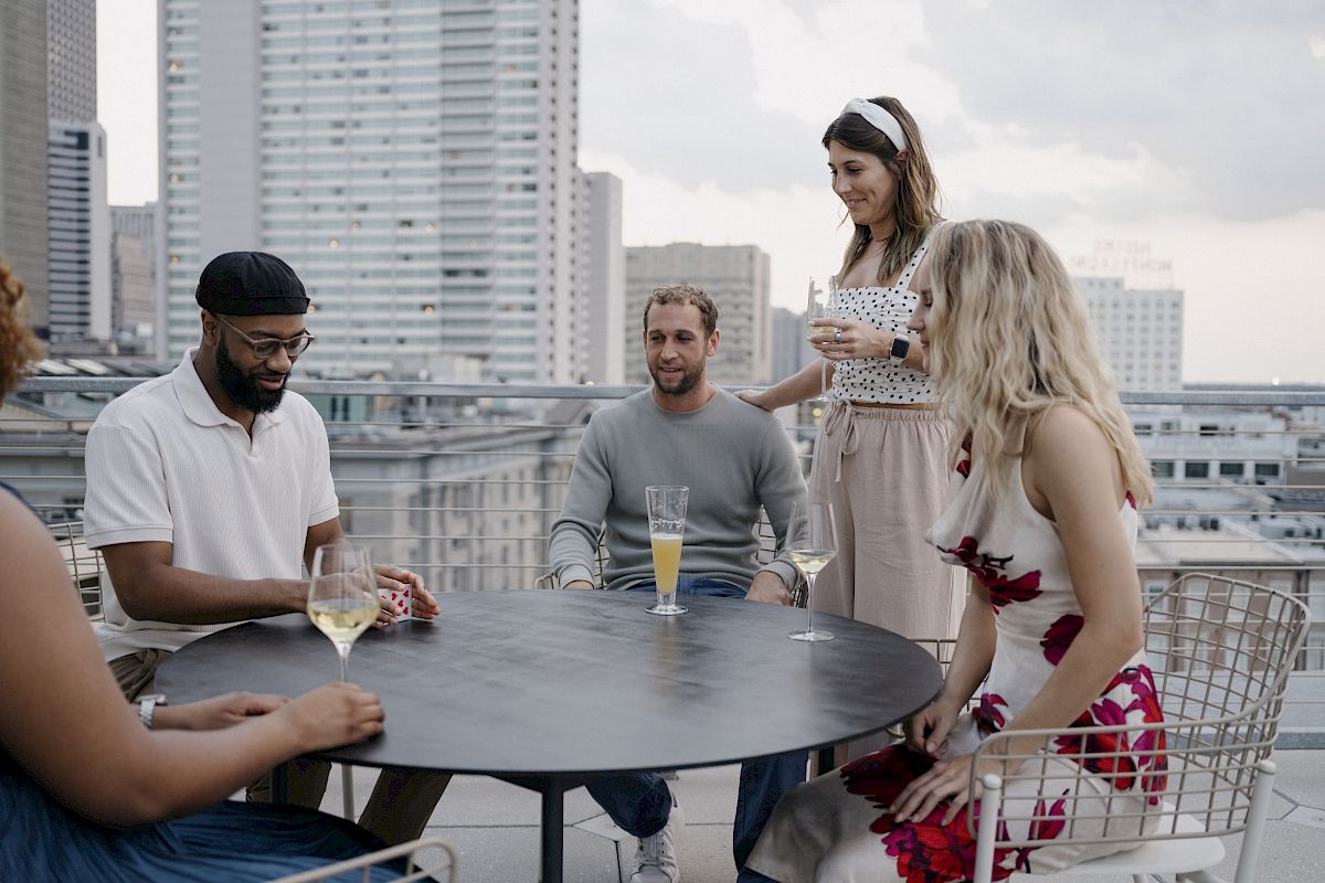 A group of five people are gathered around a table on a rooftop, socializing and enjoying drinks with a cityscape in the background.