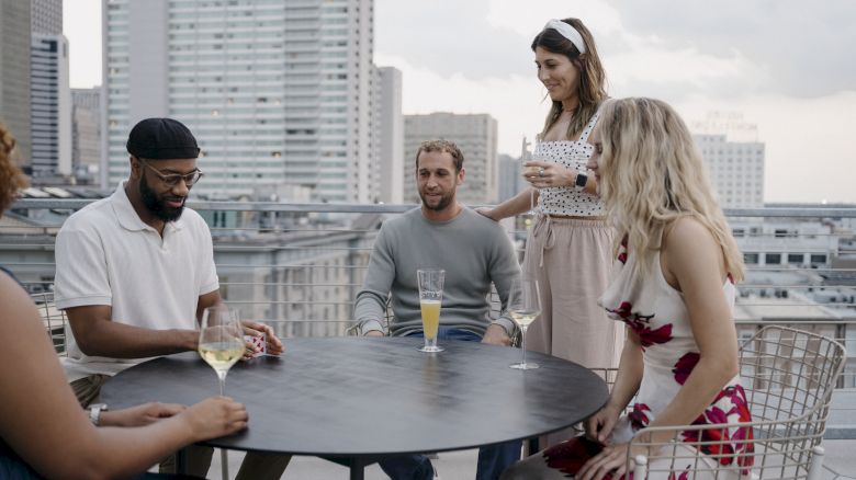 A group of five people are gathered around a table on a rooftop, socializing and enjoying drinks with a cityscape in the background.