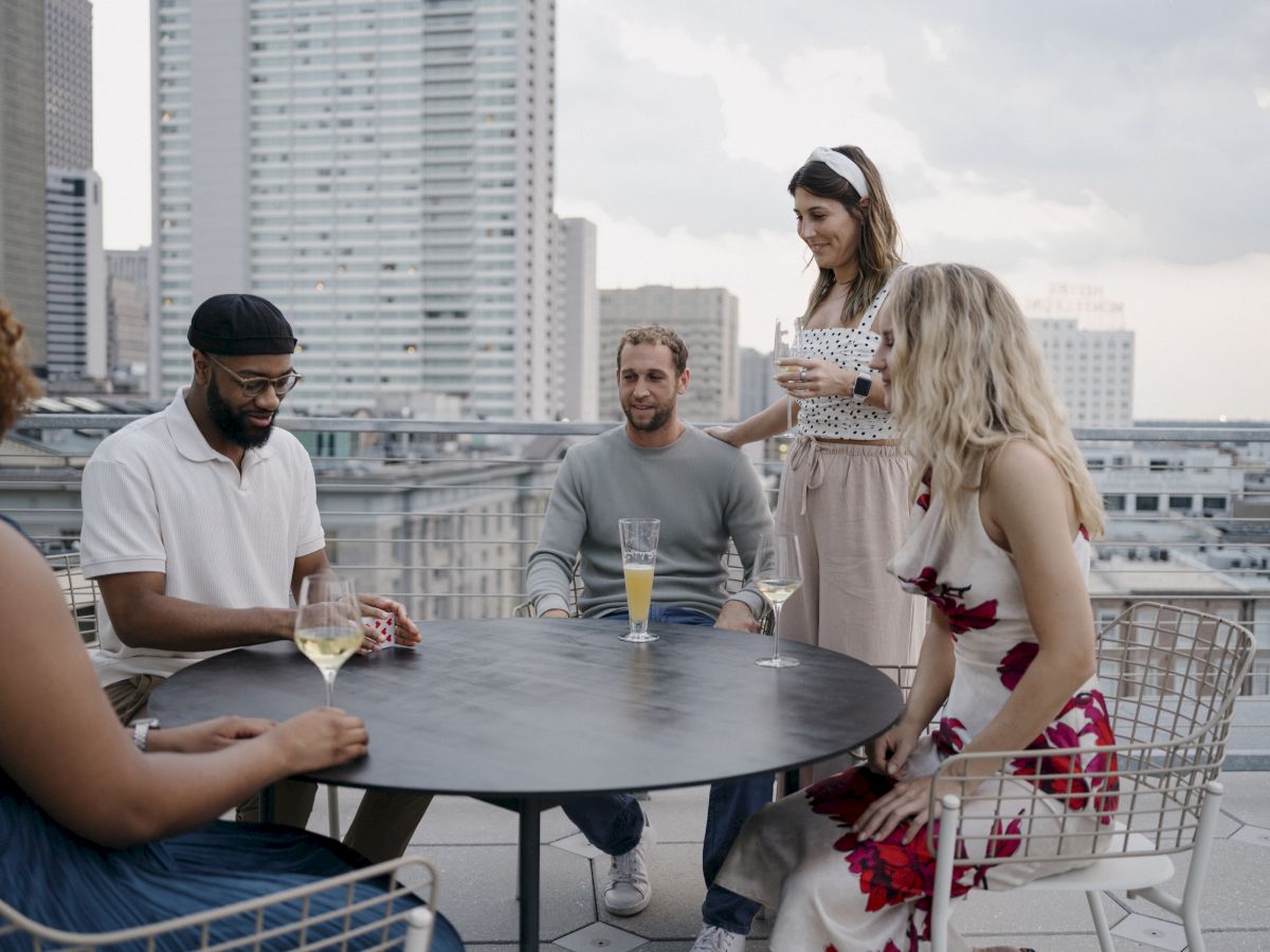A group of five people are gathered around a table on a rooftop, socializing and enjoying drinks with a cityscape in the background.