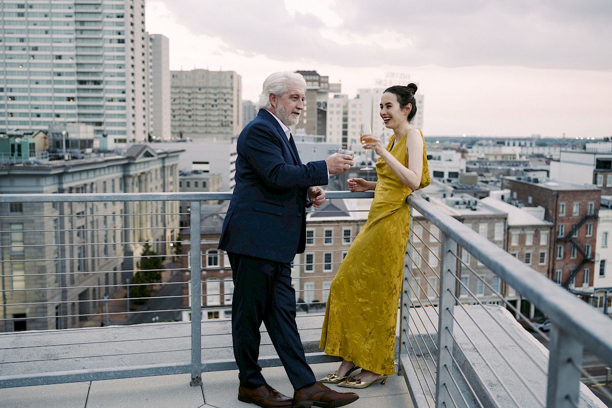 A man and a woman are standing on a rooftop, enjoying drinks with a cityscape backdrop. They appear to be engaged in conversation.