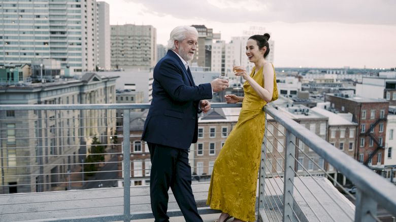 A man and a woman are standing on a rooftop, enjoying drinks with a cityscape backdrop. They appear to be engaged in conversation.