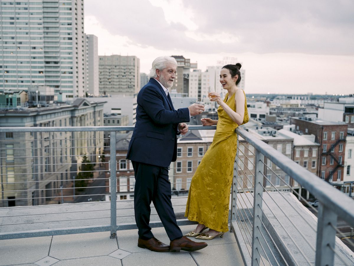 A man and a woman are standing on a rooftop, enjoying drinks with a cityscape backdrop. They appear to be engaged in conversation.