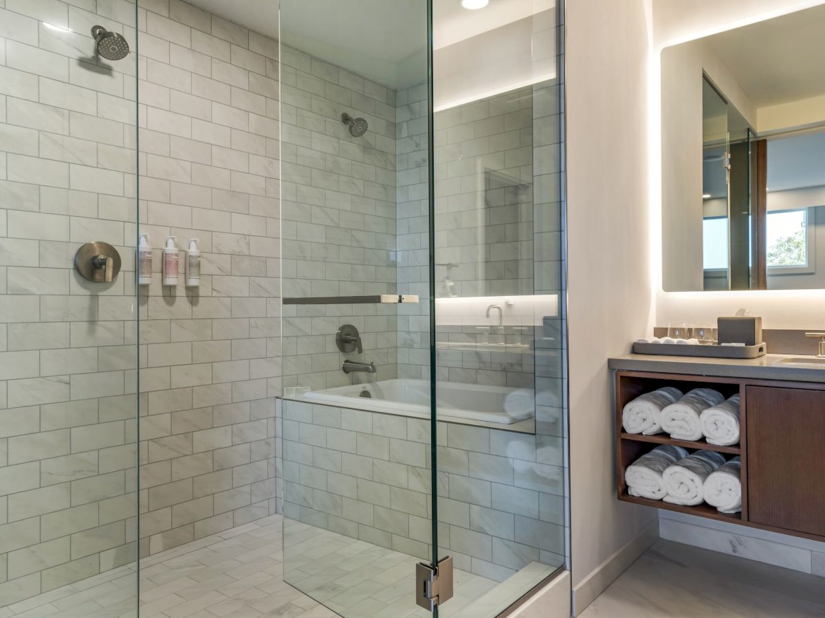 The image shows a modern bathroom with a glass-enclosed shower, a well-lit vanity, and shelves with neatly rolled towels.