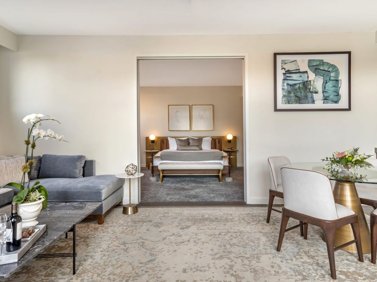 A modern living room with a gray couch, a dining area, and a view of a bedroom with a double bed in the background.
