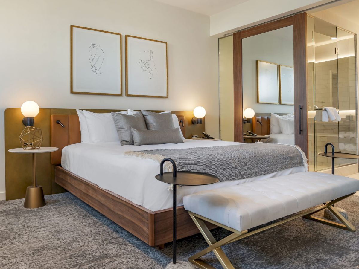 The image shows a modern bedroom with a large bed, neutral decor, two nightstands with lamps, a bench at the foot of the bed, and a glass-enclosed bathroom.
