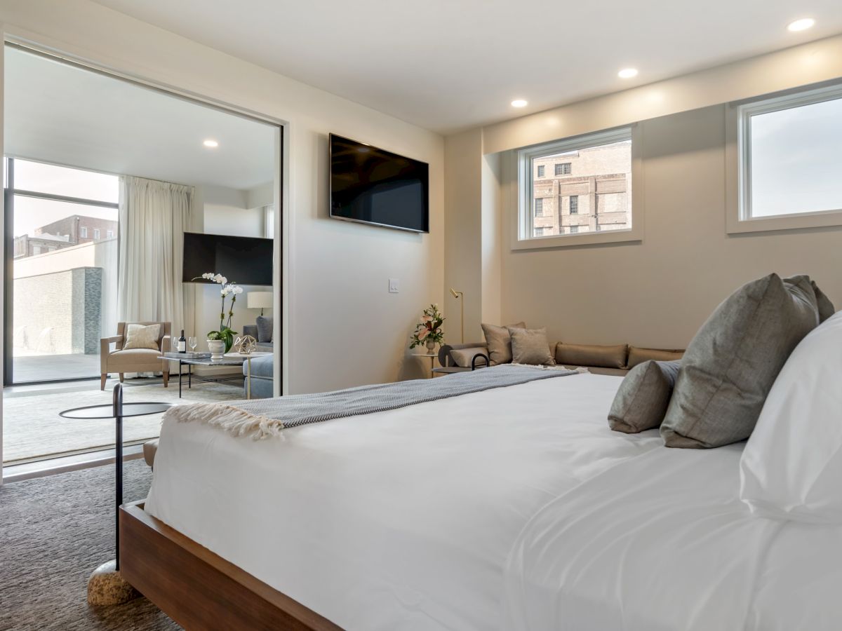 A modern, well-lit bedroom with a bed, sofa, wall-mounted TV, and two windows. An adjacent sitting area with chairs and a sliding glass door.