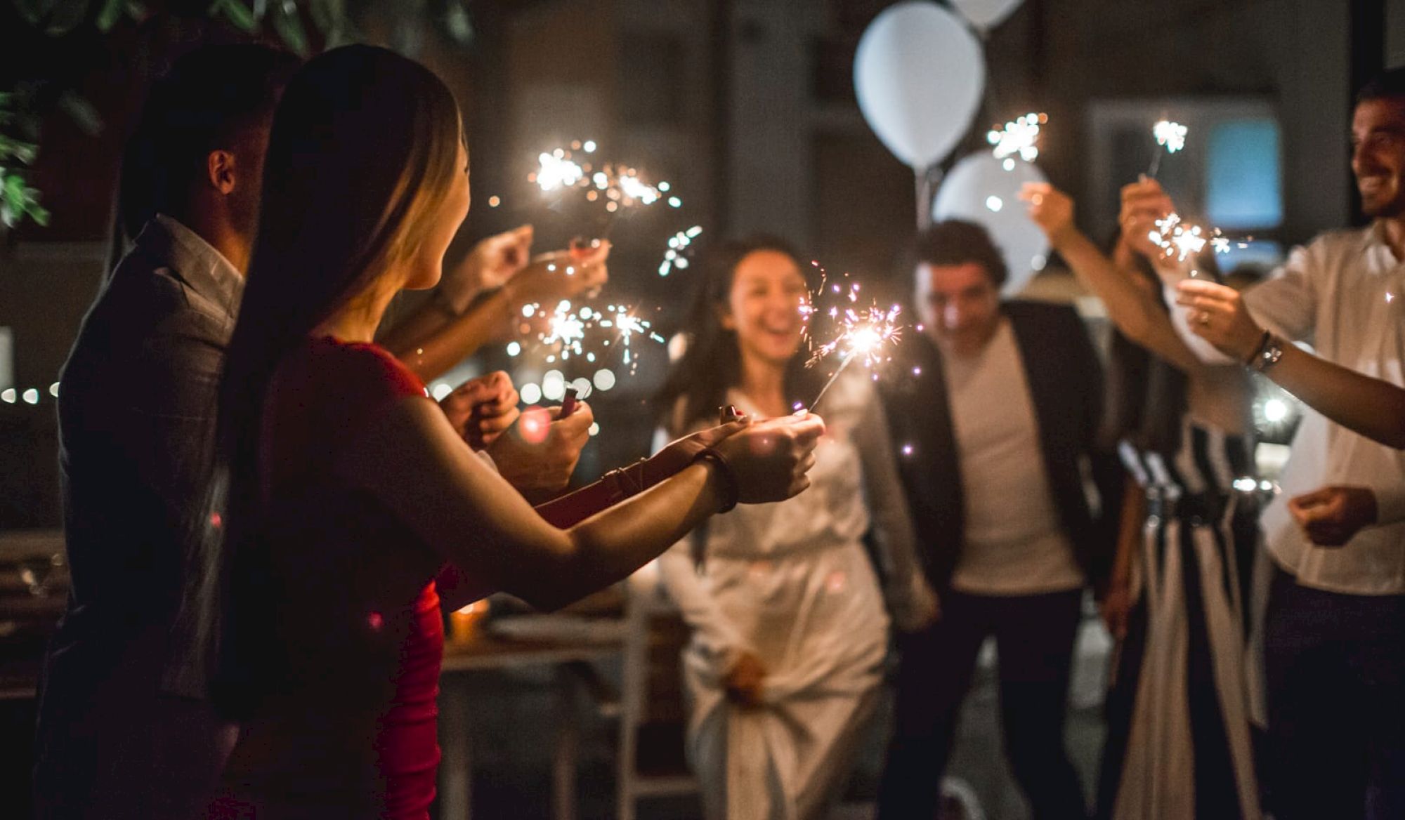 A group of people is celebrating at a party, holding and waving sparklers with smiles and balloons in the background.