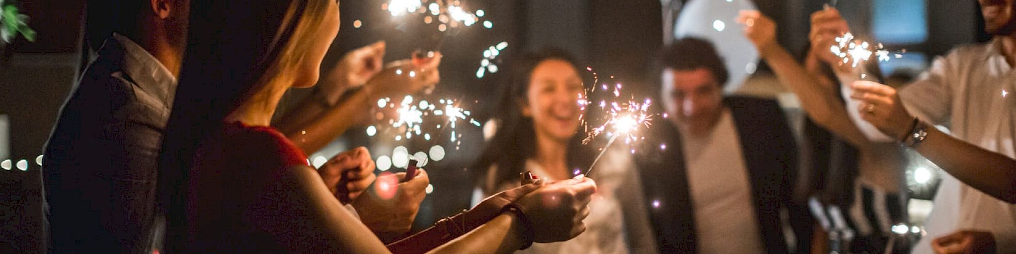 A group of people is celebrating at a party, holding and waving sparklers with smiles and balloons in the background.