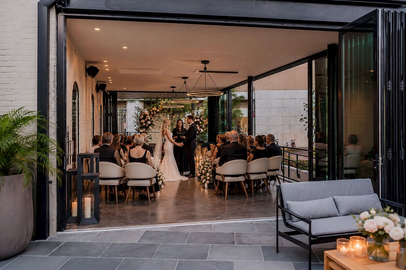 A wedding ceremony taking place in a modern lobby with glass doors, white walls, and a seating area.