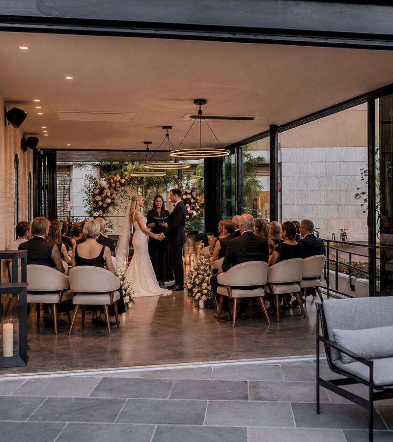 a wedding ceremony with a bride and groom and guests in a room with a floral arch