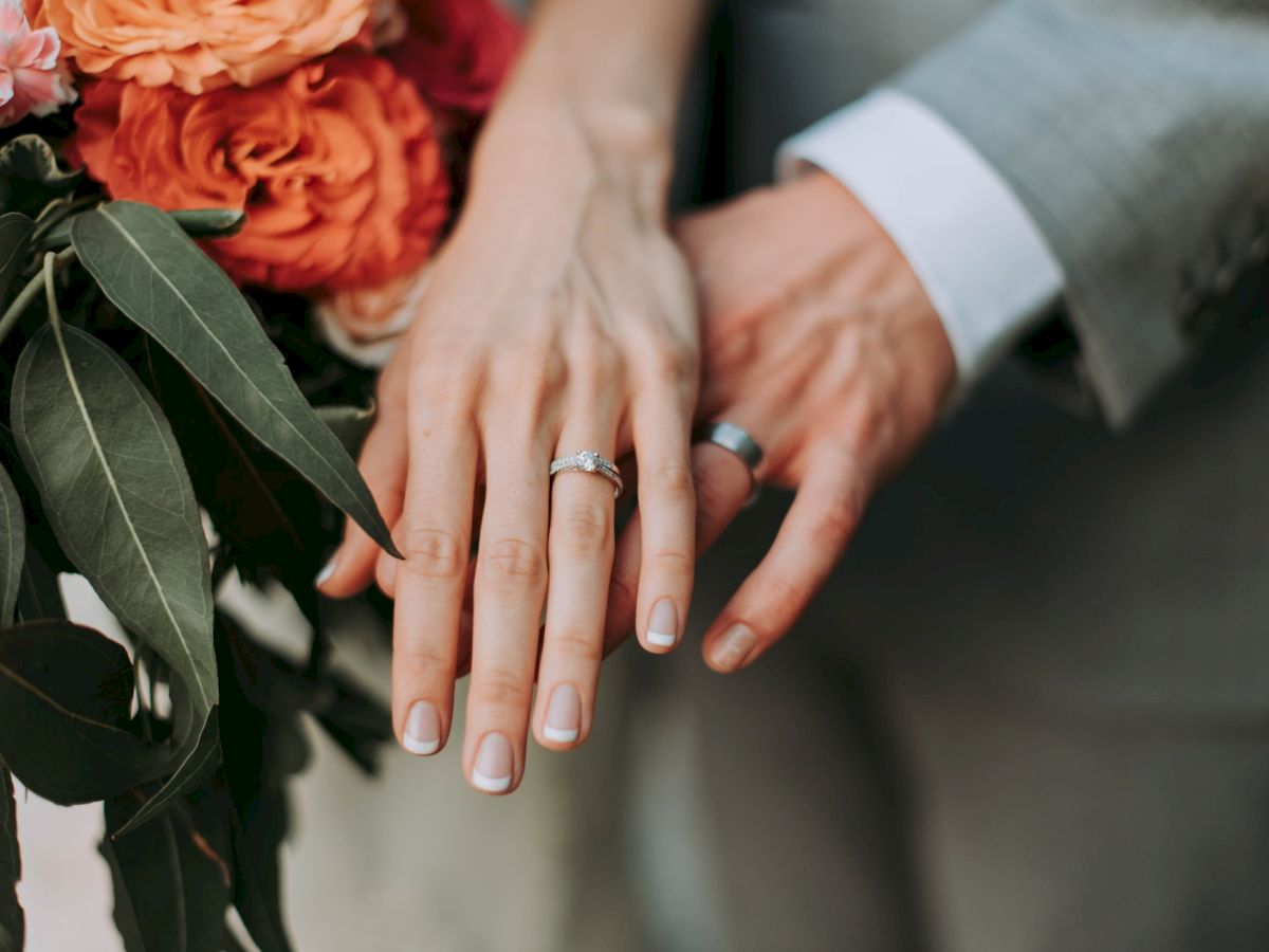 The image shows two people wearing wedding rings on their hands, with a bouquet of orange flowers and green leaves in the background.