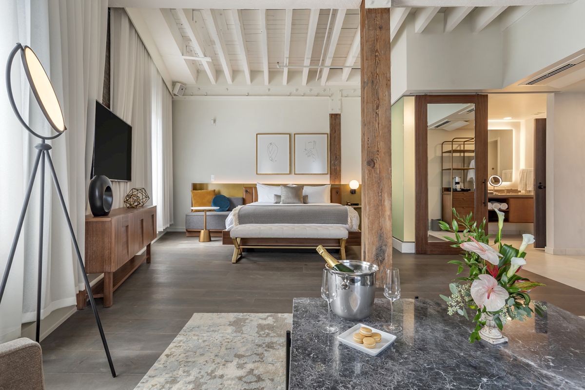 A chic hotel suite with a king bed, TV, soft lamp light, flowers, and a marble table with a champagne bucket.
