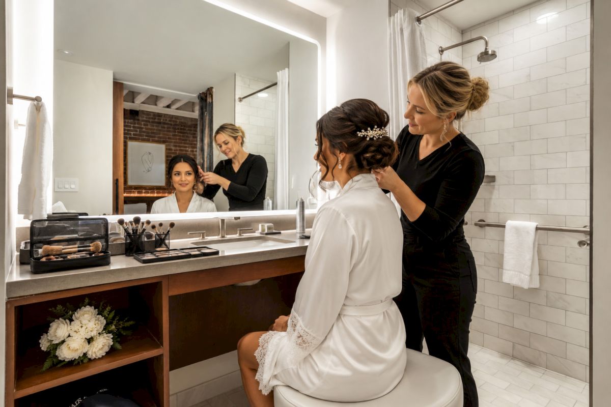 A stylist fixes a bride&rsquo;s hair and makeup in a bright salon-style bathroom, with brushes and bottles on the counter.