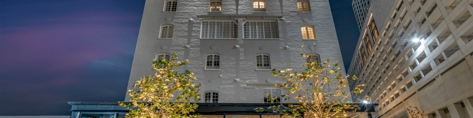 A tall illuminated building at night, featuring large windows and a ground-level restaurant with outdoor seating and decorative trees.