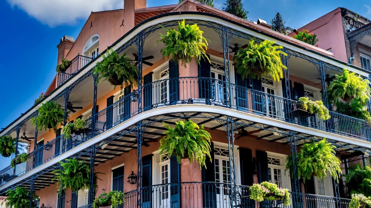 A historic building with wrought-iron balconies adorned with lush green hanging plants against a bright blue sky with scattered clouds.