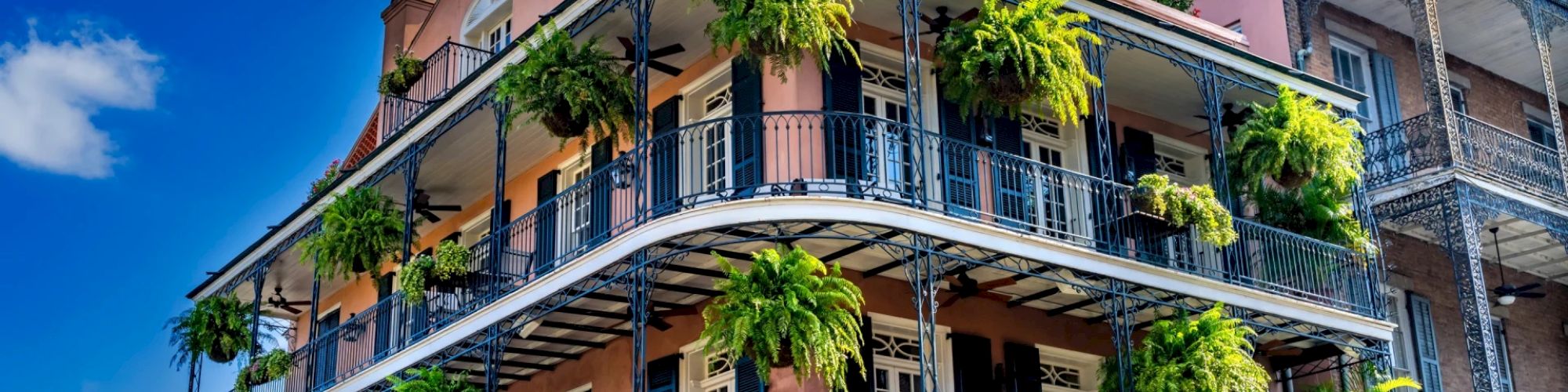 A historic building with wrought-iron balconies adorned with lush green hanging plants against a bright blue sky with scattered clouds.