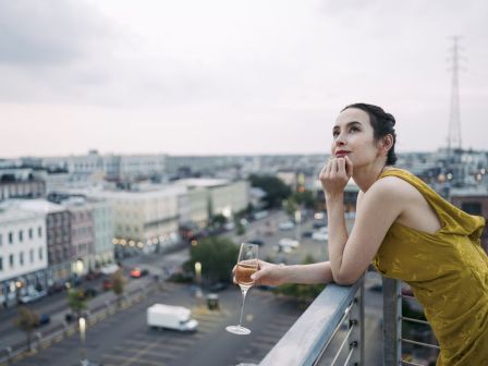 A woman in a yellow dress sips champagne on a rooftop, overlooking a city skyline at dusk.