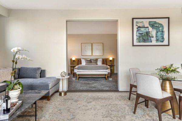 The image shows a modern, well-lit living room with a sofa, coffee table, plants, and artwork, leading to a bedroom with a neatly made bed.