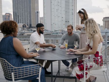 Friends play cards on a rooftop patio, laughing over drinks with city skyscrapers in the background.