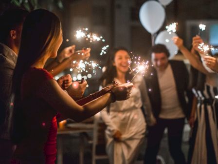 A group of people holding sparklers are gathered together at a festive event with balloons visible in the background, looking joyful and celebratory.