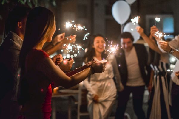 A group of people holding sparklers are gathered together at a festive event with balloons visible in the background, looking joyful and celebratory.