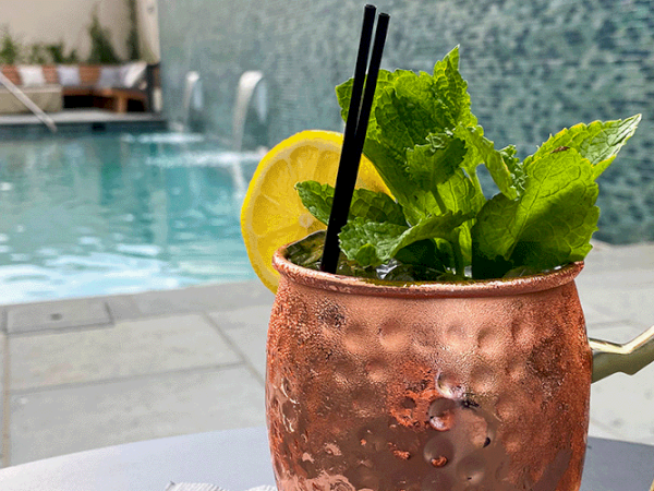 A copper mug with a drink, garnished with mint and lemon, sits by a poolside with lounge chairs and water features in the background.