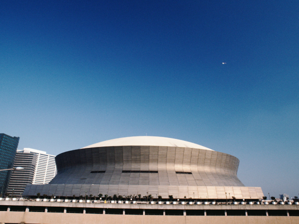 The image shows a large, dome-shaped building with a clear blue sky in the background. Some modern buildings are visible to the left.