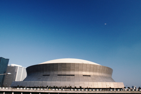 The image shows a large, dome-shaped building with a clear blue sky in the background. Some modern buildings are visible to the left.