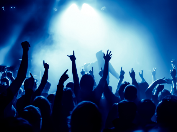 A crowd of people at a concert, raising their hands and enjoying the music. The stage is lit with blue lighting and a bright spotlight.