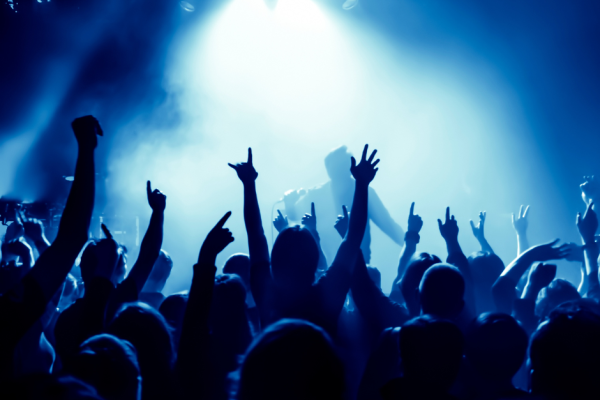 A crowd of people at a concert, raising their hands and enjoying the music. The stage is lit with blue lighting and a bright spotlight.