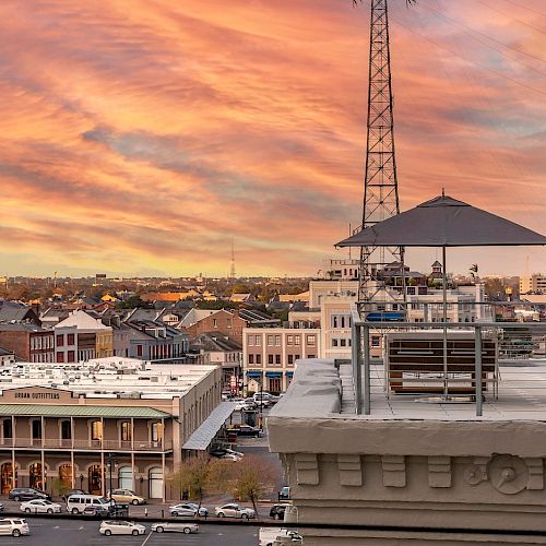 Cityscape at sunset with buildings, a church spire, and a viewing deck with seating and an umbrella in the foreground.