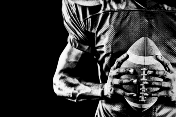 A black and white image of an American football player holding a football tightly against their chest, wearing a textured jersey.